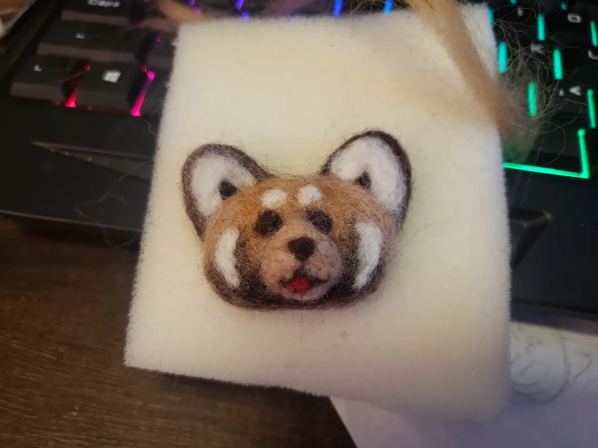 A close-up photo shows a small, needle-felted red panda head crafted from brown, white, and black wool fibers, resting on a…