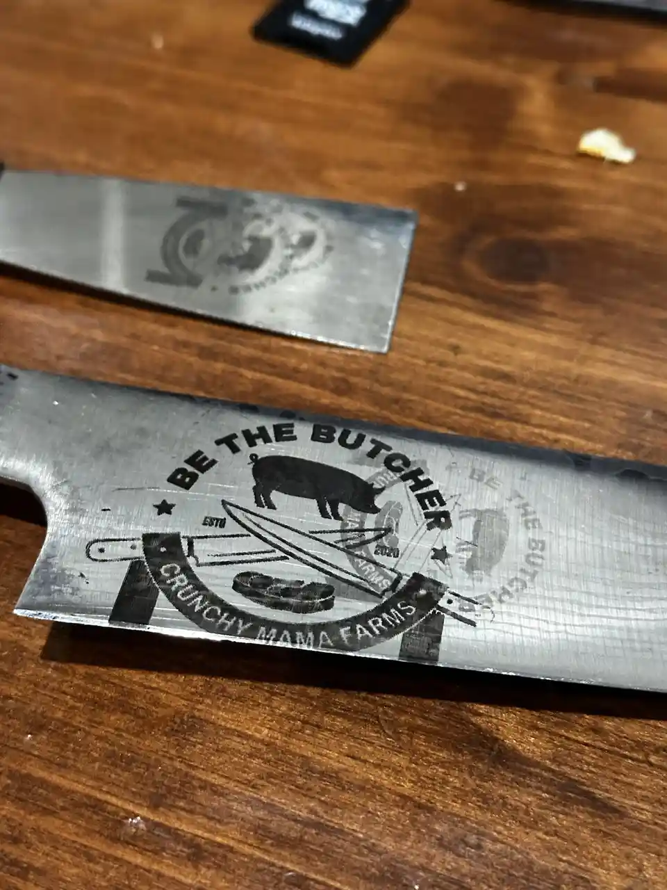 A close-up photo shows two metal cleavers resting on a wooden table. The cleaver in the foreground has an engraved logo…