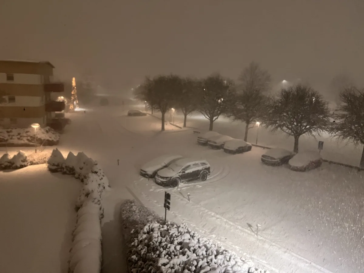 A nighttime photo of a snowy residential parking lot with cars buried under snow, streetlights casting a glow through…
