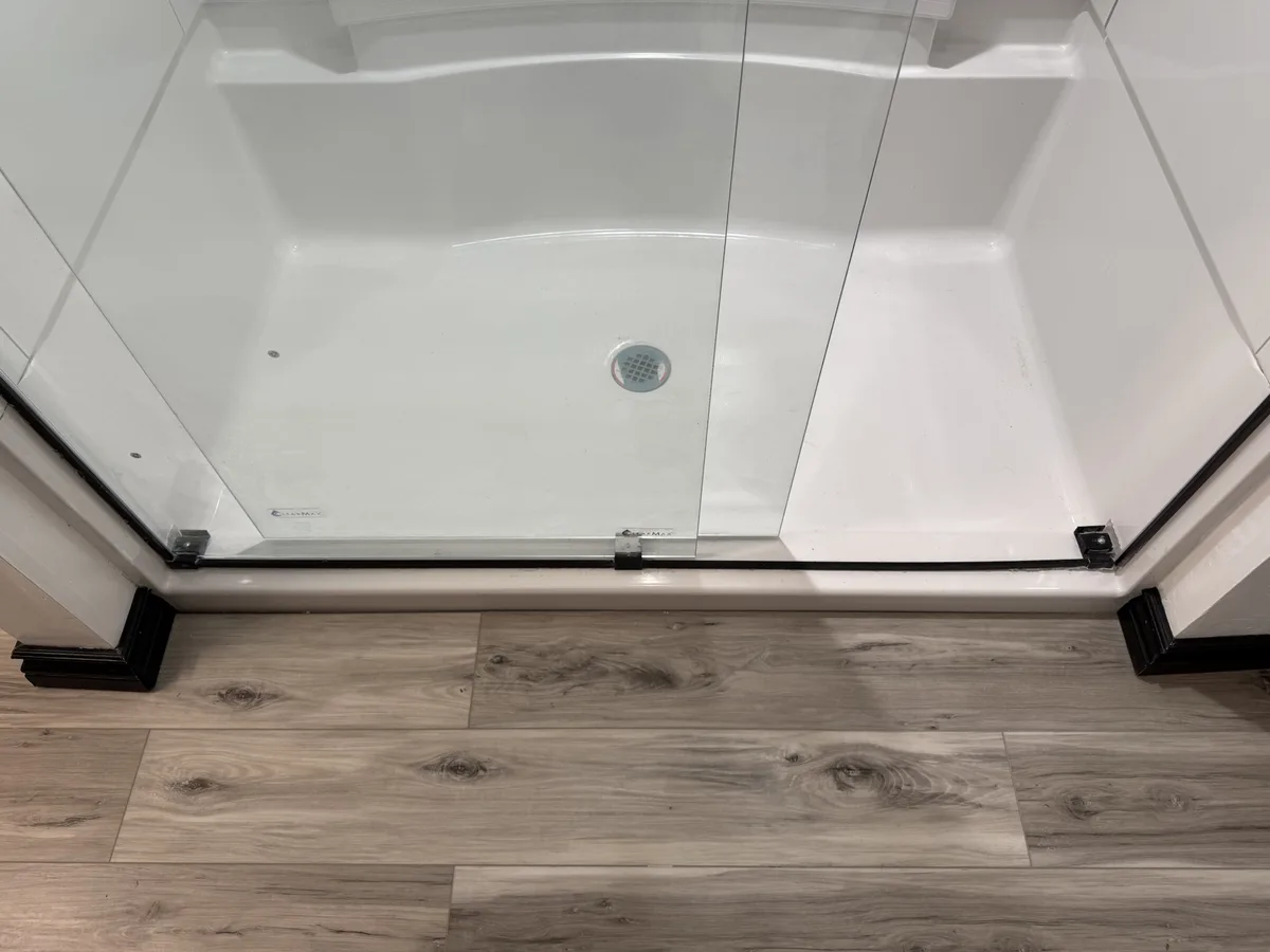 A top-down view of a clean, white, empty walk-in shower with clear glass doors and black trim, set against a light…