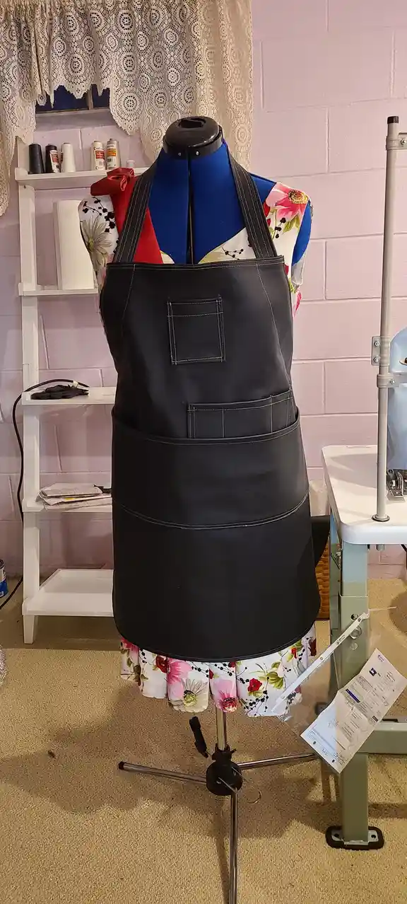 A mannequin in a sewing room wears a black apron over a floral dress, standing next to a sewing machine and shelves with…