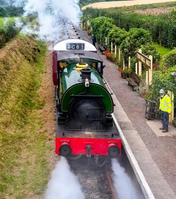 A high-angle photo shows a green and red steam locomotive, numbered 2061, emitting smoke as it approaches a station…