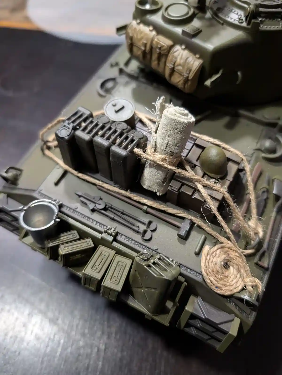 A close-up photo of a detailed green military tank model, showing its top surface loaded with miniature equipment including…