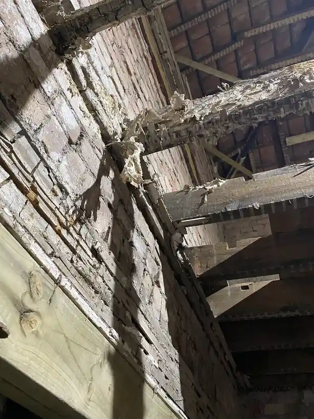 A low-angle shot of a severely deteriorated interior with crumbling brick walls, rotting wooden beams, and exposed…