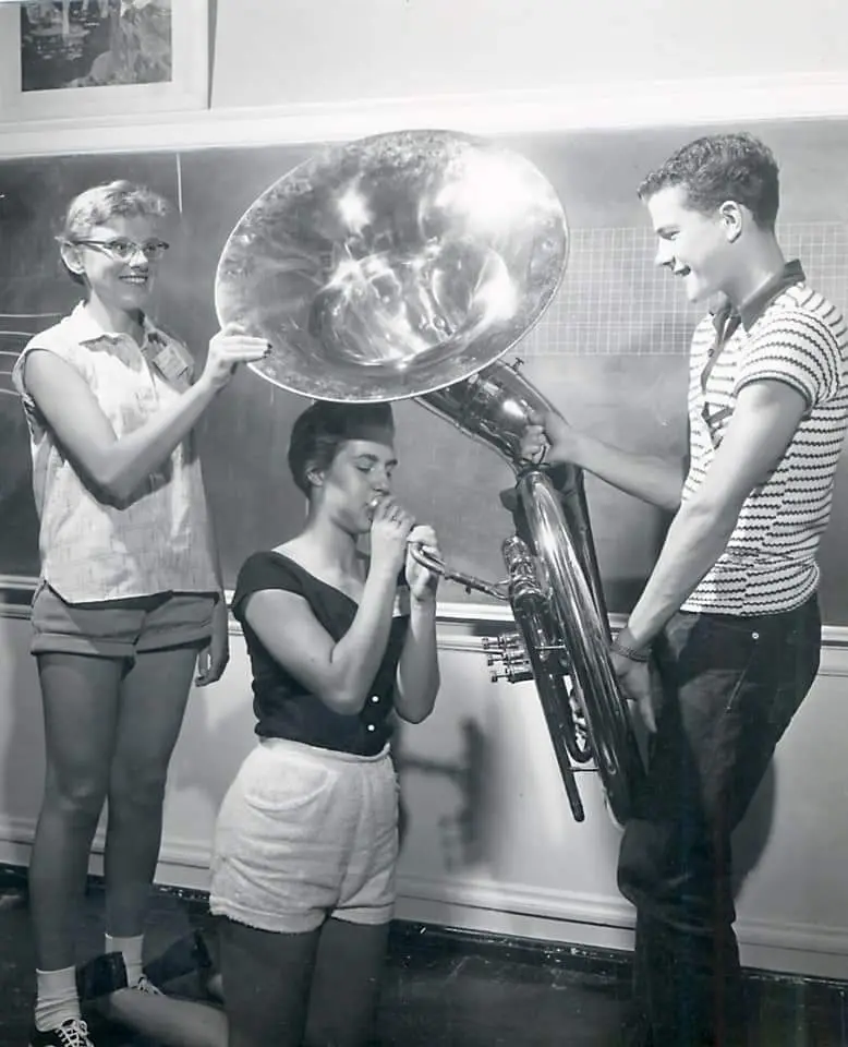 A vintage black-and-white photo shows three young people with a tuba. A woman kneels and plays the instrument while a man…