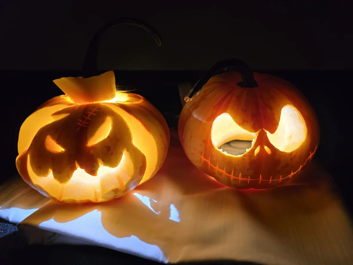 Two glowing jack-o'-lanterns sit side by side on a cloth in a dark room; the one on the left has a stitched, bat-like face…