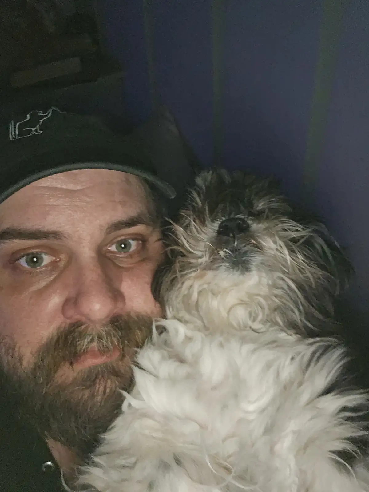 A man with a beard and a black cap is taking a selfie while holding a small, fluffy, white-and-gray dog that is resting its…