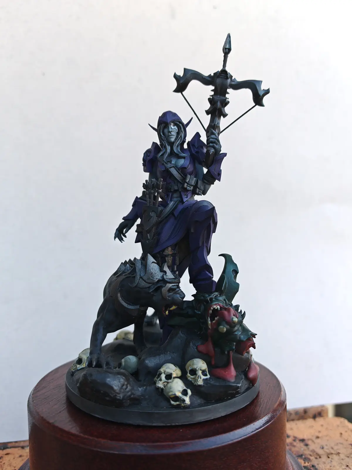 A detailed figurine of a fantasy archer with dark purple armor and a large ornate bow stands atop a base with skulls and…