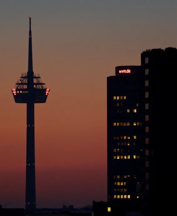 A dusk photo shows the silhouette of the TV tower in Düsseldorf next to a dark building with 'wvm.de' lit in red on its…