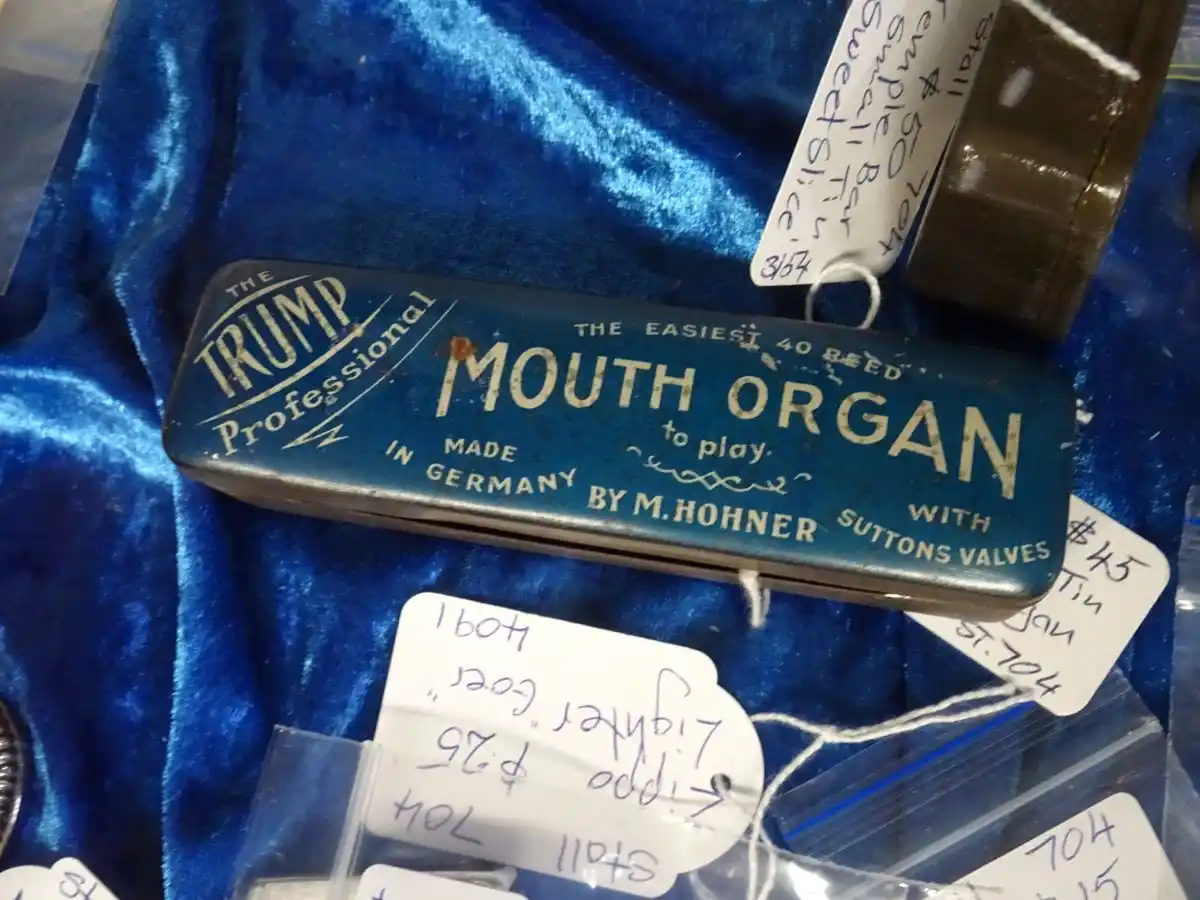 A vintage blue tin case for a 'Trump Professional Mouth Organ' is displayed on a blue velvet surface, surrounded by…