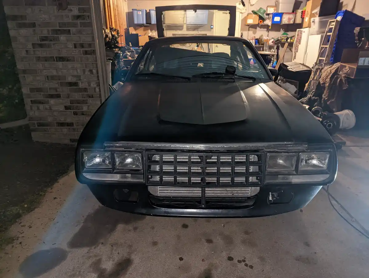 A black classic car is parked in a cluttered garage with its hood open, revealing a large aftermarket radiator installed in…