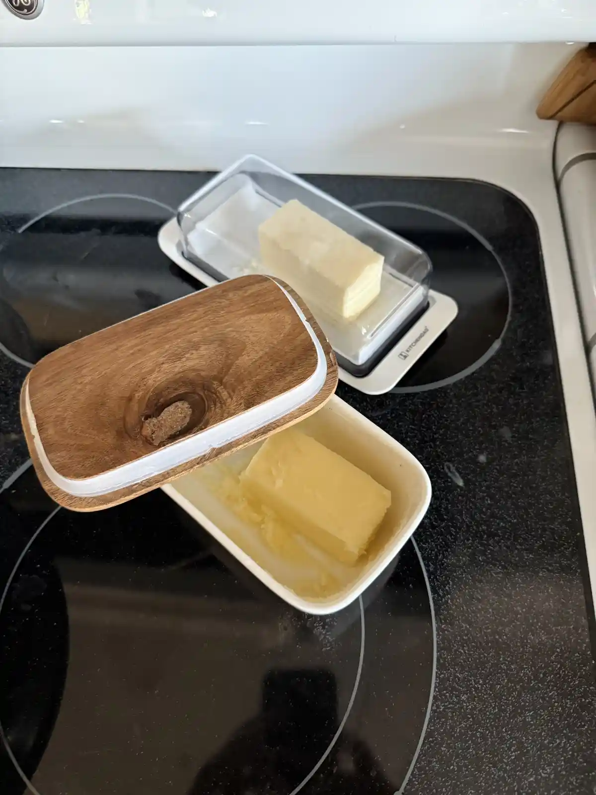 Two butter containers sit on a black electric stovetop; one has a wooden lid and contains a partially melted stick of…