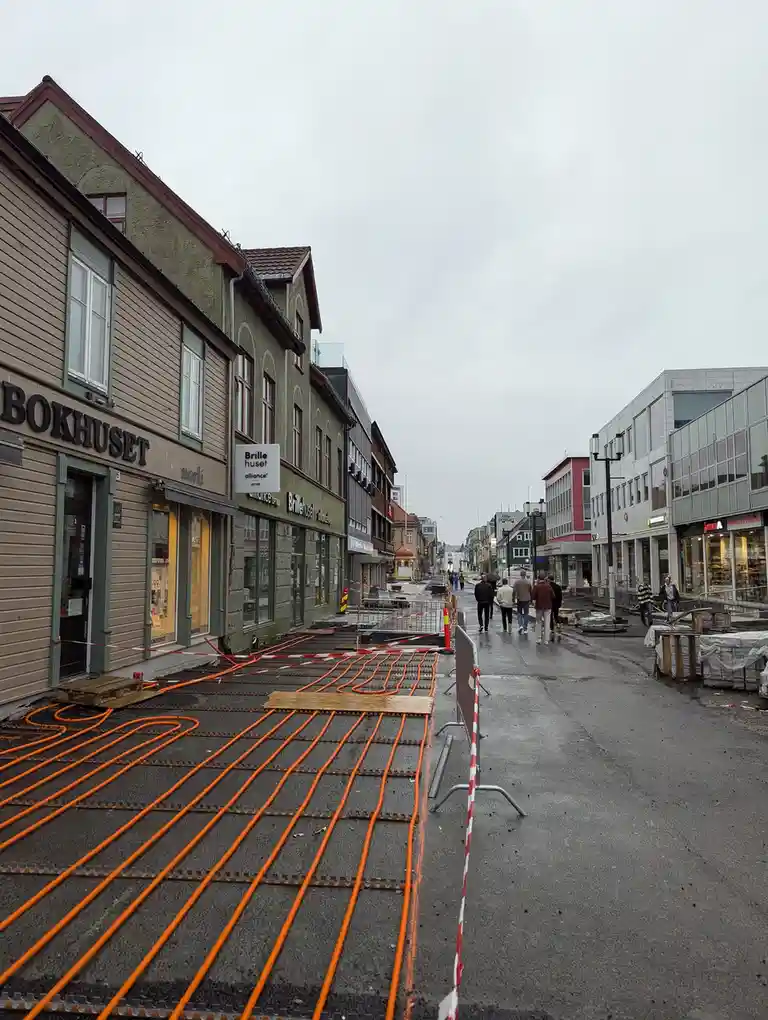A street scene shows orange pipes laid out in a grid pattern on the asphalt, surrounded by red-and-white barriers, with…