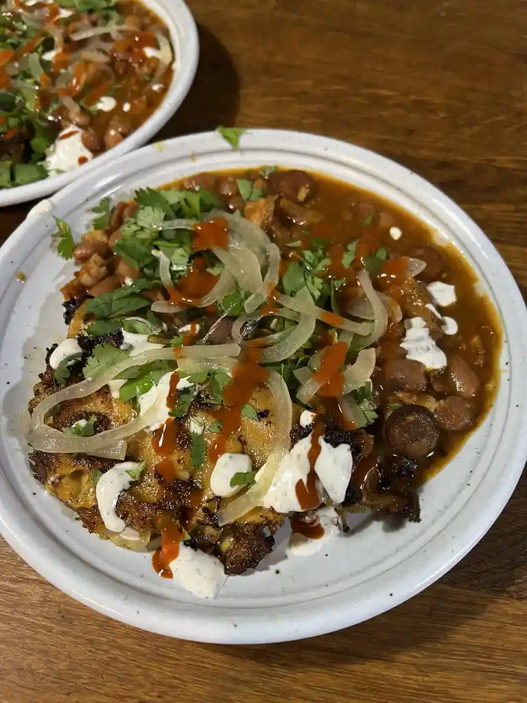 A close-up photo shows a white bowl filled with a messy, saucy dish that resembles a taco or nacho, topped with cilantro…