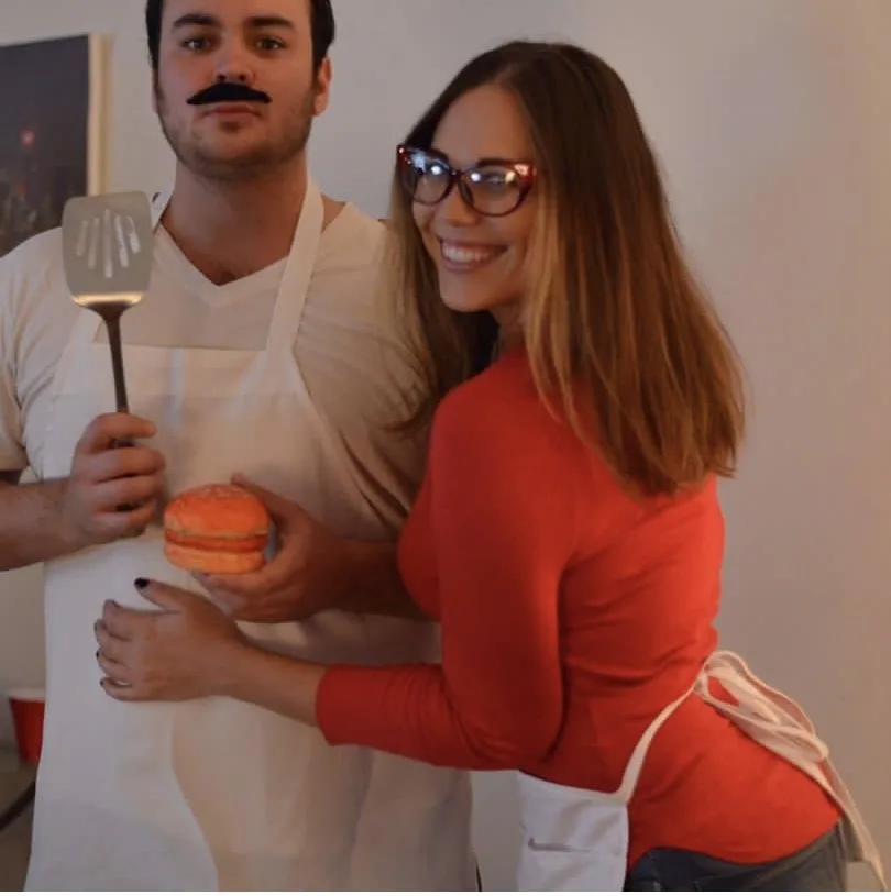 A man wearing a fake mustache and apron holds a spatula and a macaron, while a smiling woman in glasses and an apron holds…