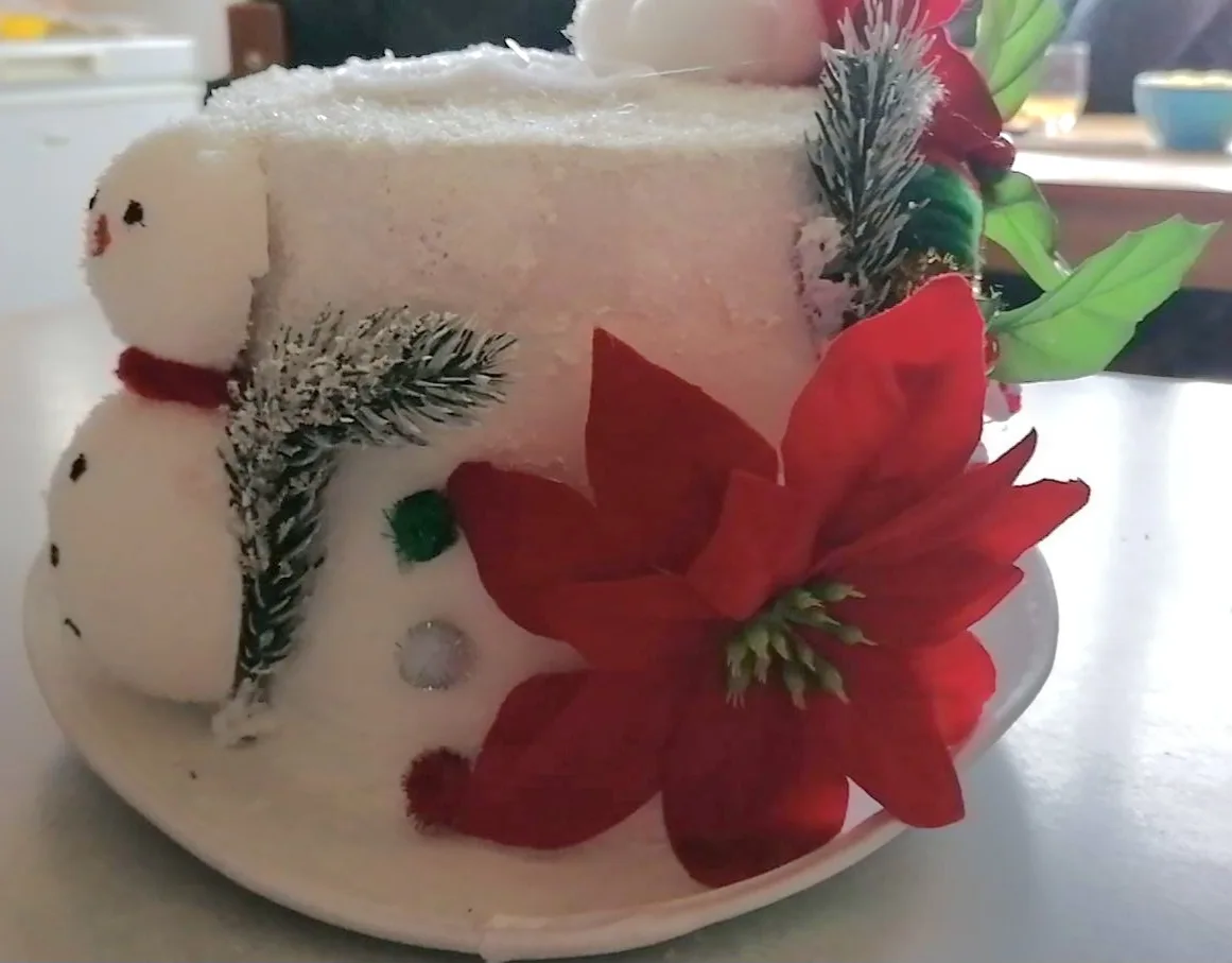 A white frosted cake decorated to resemble a snowman, featuring two stacked snowball tiers with black dot eyes and a red…