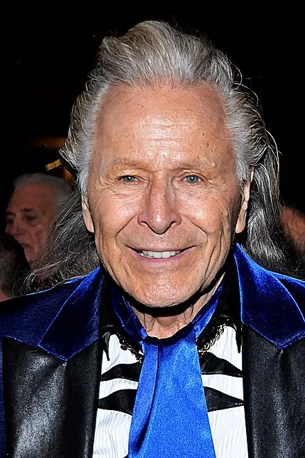 A close-up photo of an older man with long gray hair, wearing a black leather jacket with a blue velvet collar, a…