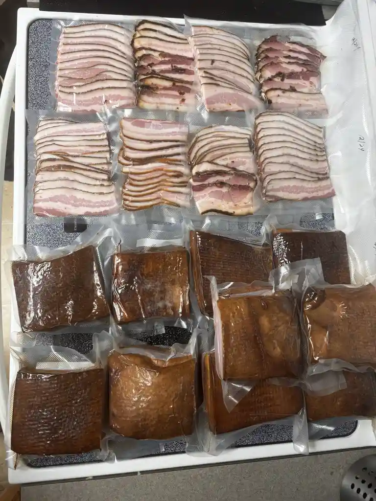 An overhead shot shows a white tray filled with multiple vacuum-sealed packages of bacon. The top rows contain raw bacon…
