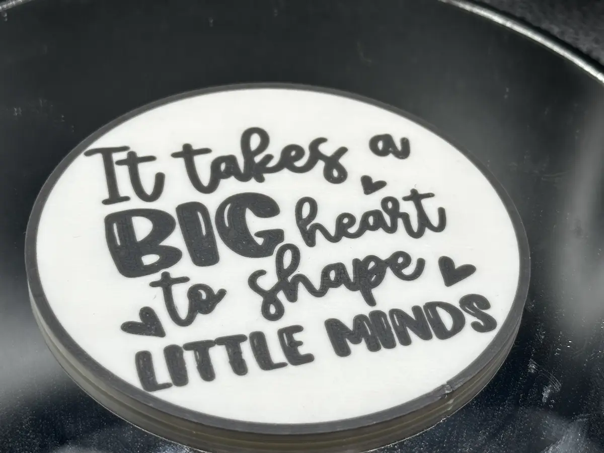 A close-up of a white oval coaster with black text that reads 'It takes a BIG heart to shape LITTLE MINDS', with small heart…