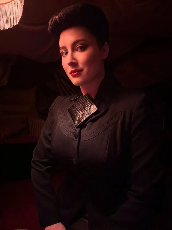 A woman with a dark, slicked-back hairstyle and red lipstick stares directly at the camera with a serious expression. She…