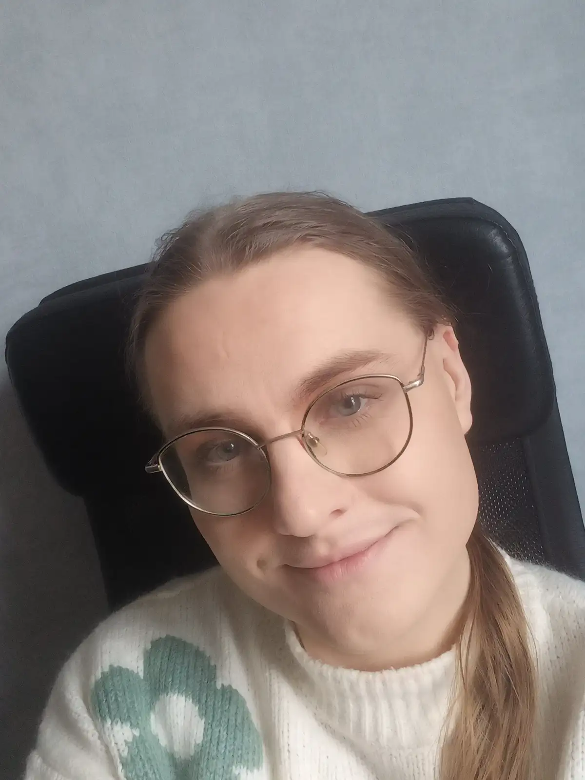 A person with light brown hair, wearing round glasses and a white sweater with a green floral pattern, leans back in a black…