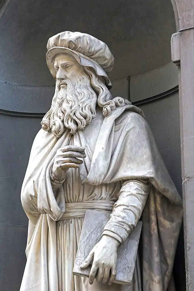 A marble statue of Leonardo da Vinci, depicted with a long beard, wearing a hat and robe, holding a book or sketchpad. The…