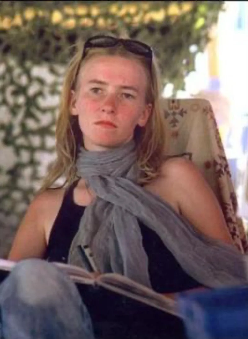 A woman with blonde hair and sunglasses perched on her head sits in a chair, wearing a grey scarf and black top, holding an…