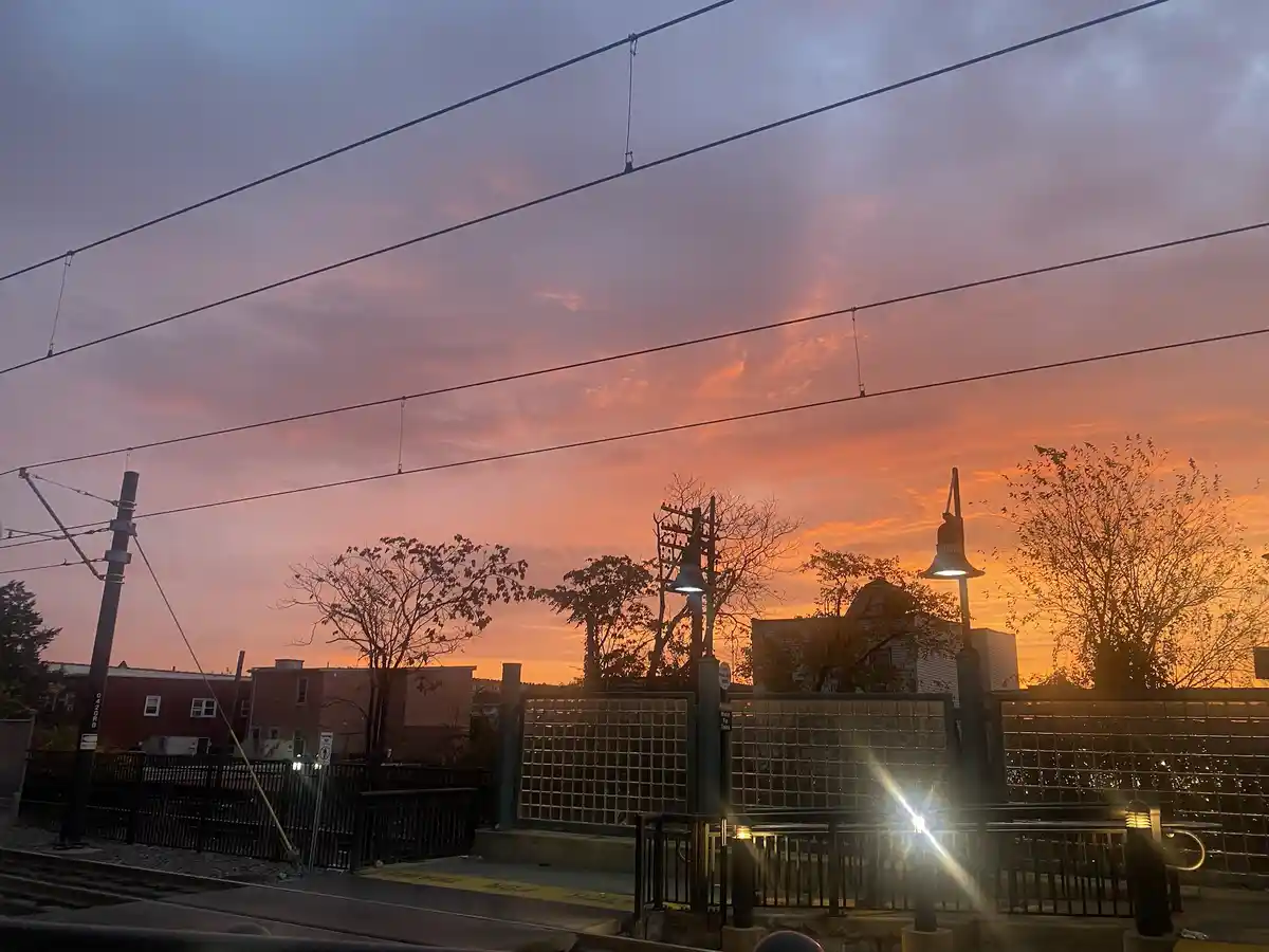 A photograph of a train station platform at sunset, with overhead power lines, a glass-block fence, and trees silhouetted…
