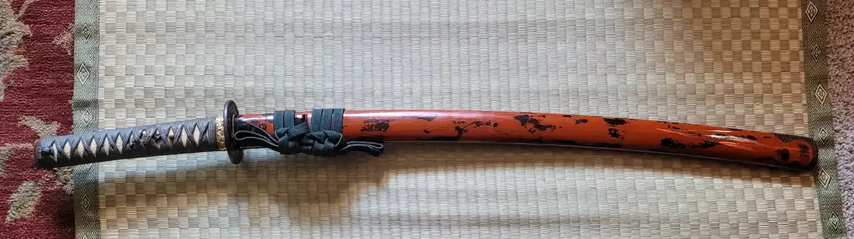 A traditional Japanese katana sword, fully sheathed in a red lacquered scabbard with black chipping, lies horizontally on a…