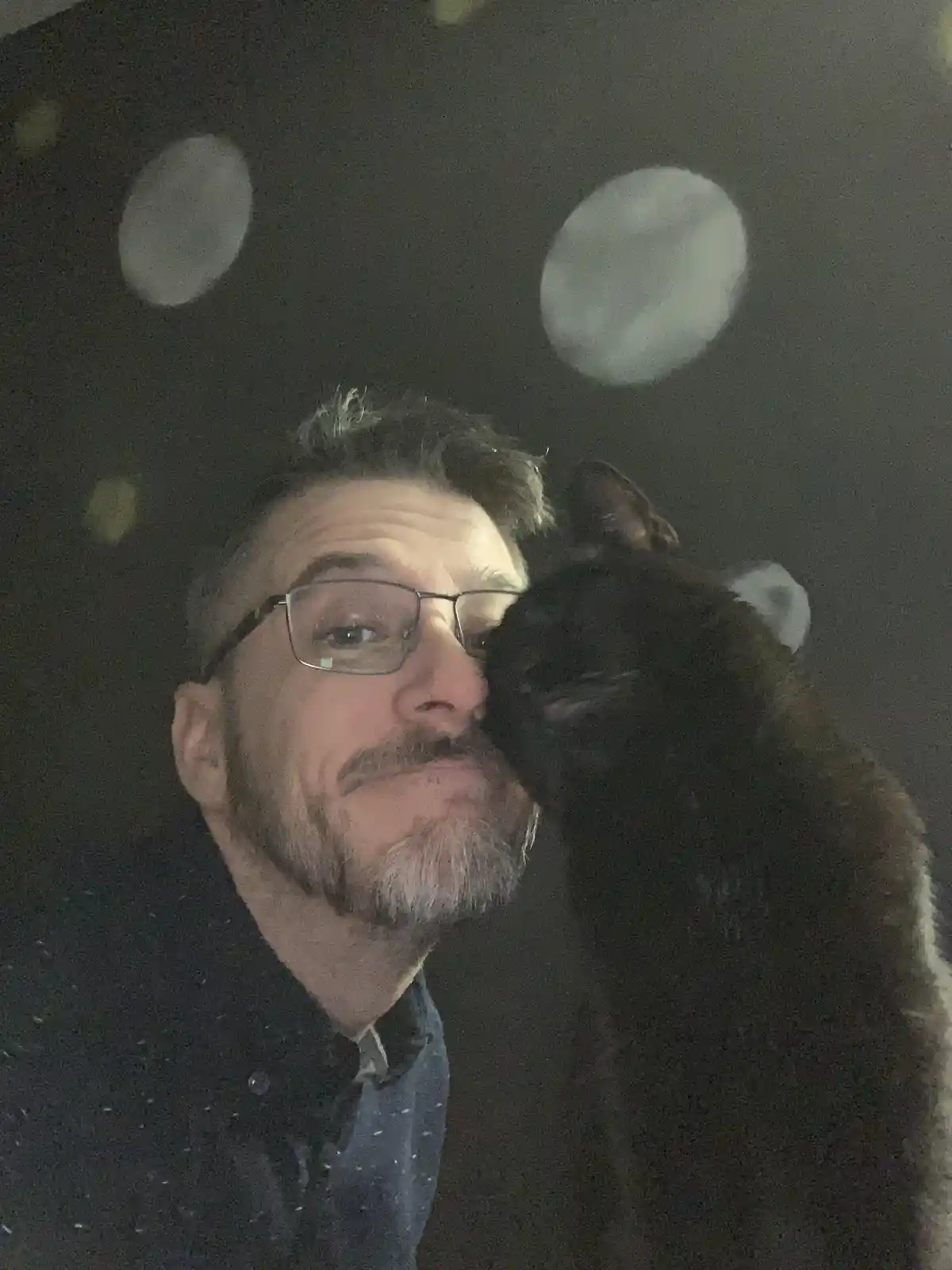 A man with glasses and a beard is taking a selfie with a black cat that is nuzzling his cheek. The background is dark with…