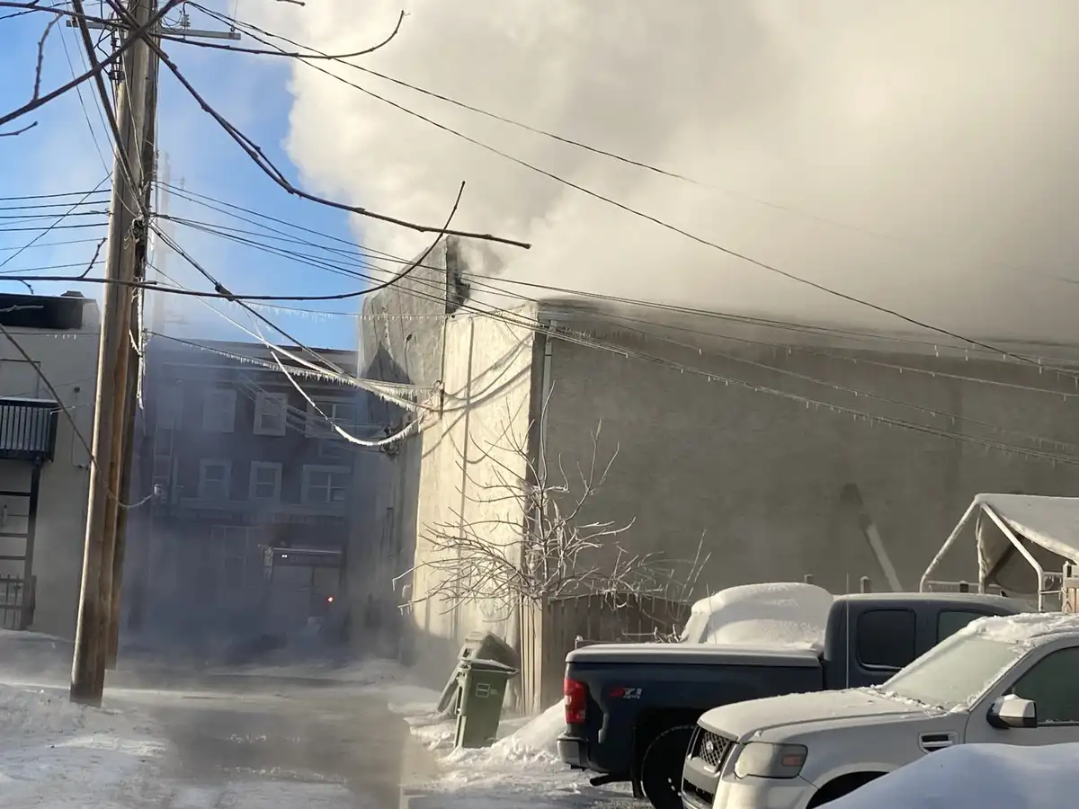 A snowy urban street scene shows steam billowing from a building's roof, with snow-covered cars and icicles hanging from…