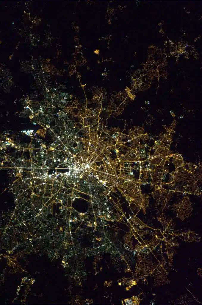 a satellite night photo of a sprawling city illuminated by countless yellow and white lights against a black background…