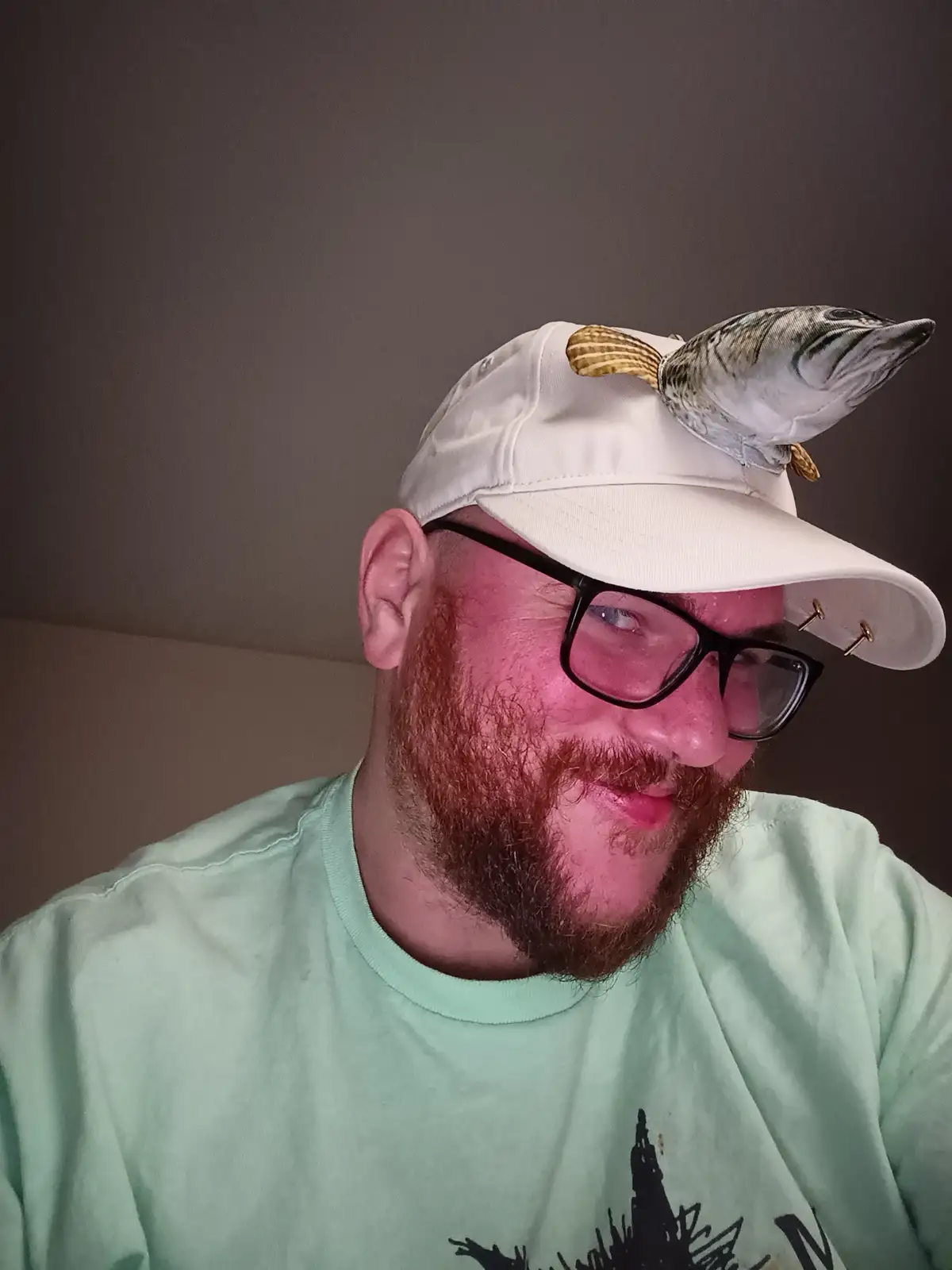 A man with a red beard and black-rimmed glasses wears a white baseball cap that has a fish-shaped horn attached to the…