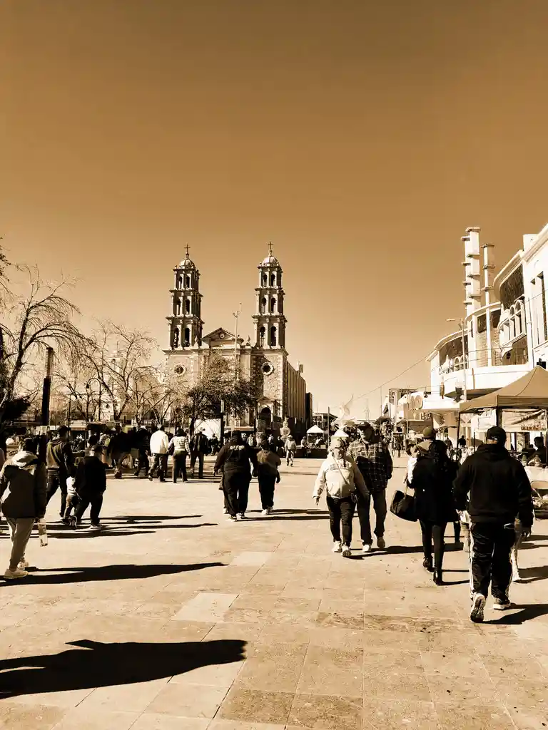 A sepia-toned photo shows a busy public square with many people walking away from the camera, toward a large church with two…