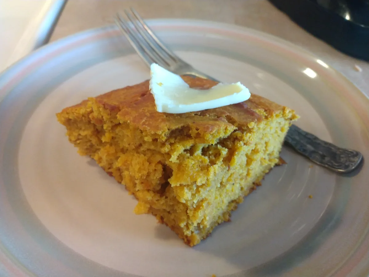 A square slice of golden-brown cornbread sits on a light-colored plate with concentric pastel stripes, topped with a pat of…