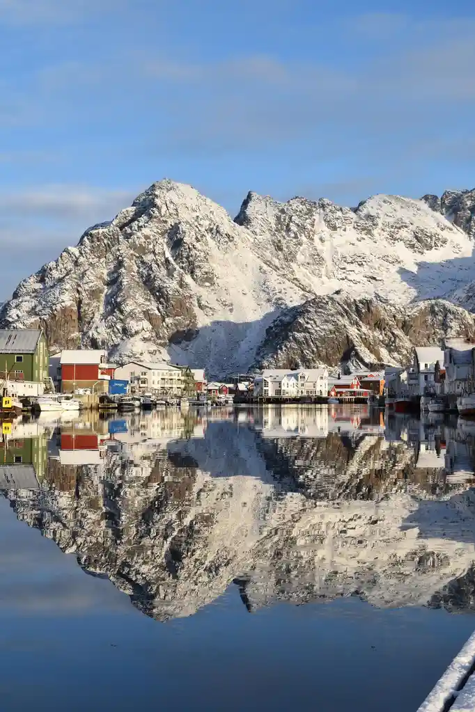 A serene winter landscape shows colorful houses and snow-covered mountains reflected perfectly in calm water. The overlaid…