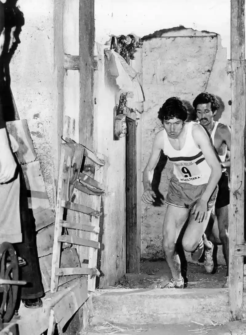 A black-and-white photo shows two male runners in vintage athletic gear emerging from a dilapidated wooden doorway. The lead…