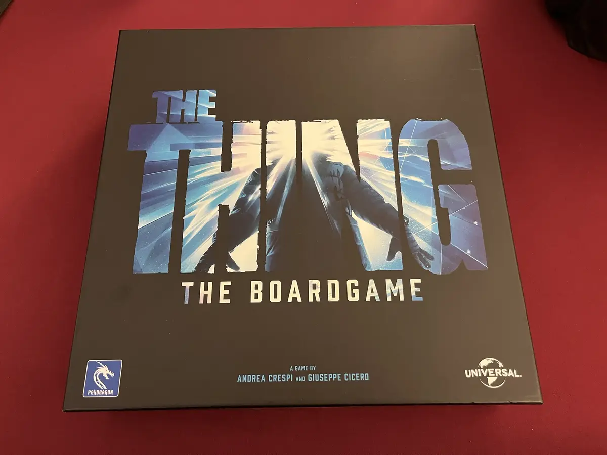 A top-down photo of the 'The Thing: The Boardgame' box, featuring a dark cover with the title in large, stylized letters…