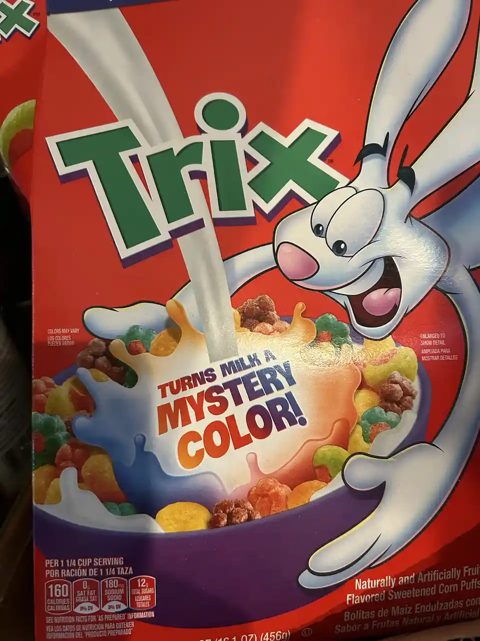 A close-up of a Trix cereal box shows the white rabbit mascot joyfully pouring milk into a bowl of colorful cereal. The milk…