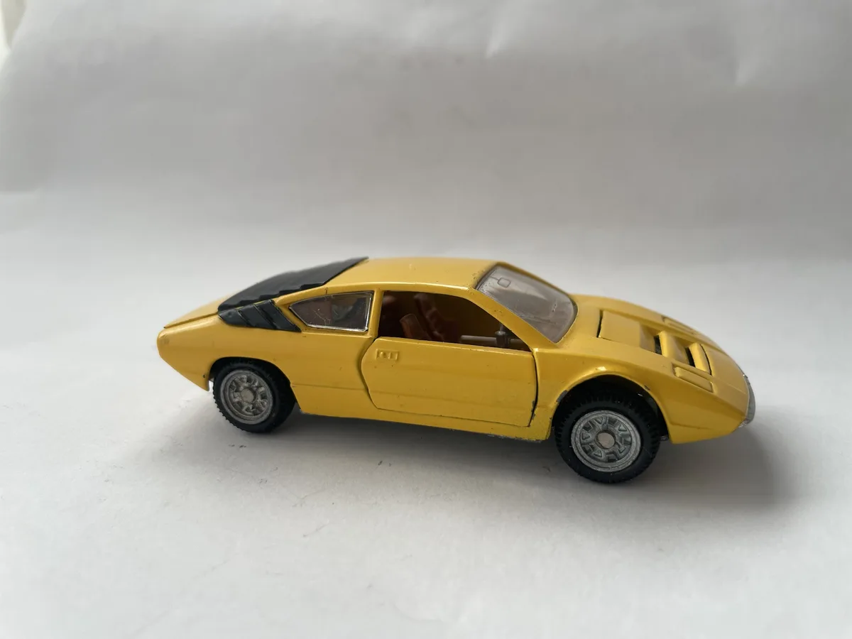 A yellow toy car with black trim and silver wheels sits on a plain white surface. The car's door is open, revealing a brown…