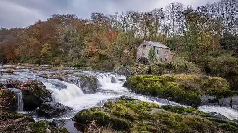 A scenic photo of a stone watermill beside a rushing river with mossy rocks and autumn trees under a cloudy sky. The…