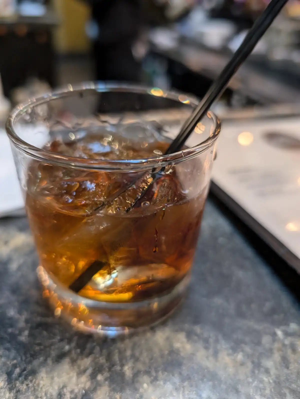 A close-up photo shows a glass of dark amber-colored cocktail with large ice cubes and a black straw, sitting on a dark…