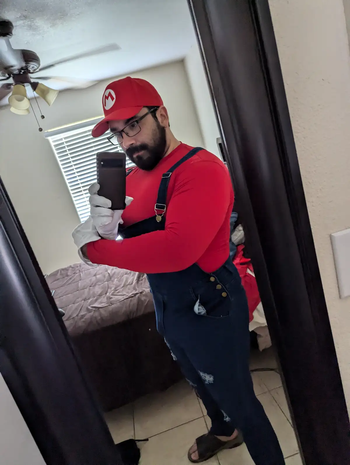 A man with a beard and glasses takes a mirror selfie while dressed in a Mario costume, wearing a red hat with an 'M' logo, a…