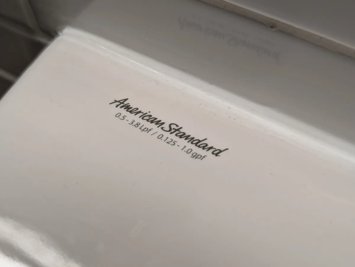A close-up photo of the bottom of a white porcelain toilet tank showing the brand name 'American Standard' and its water…