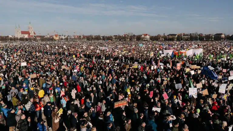 A vast crowd of people fills a large public square, holding up signs and banners under a clear sky, with a prominent church…