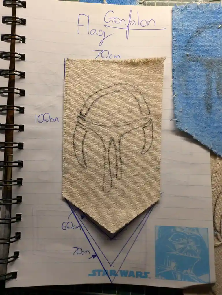 A beige fabric flag with a crude pencil sketch of a Mandalorian helmet is laid on a lined notebook. Handwritten measurements…