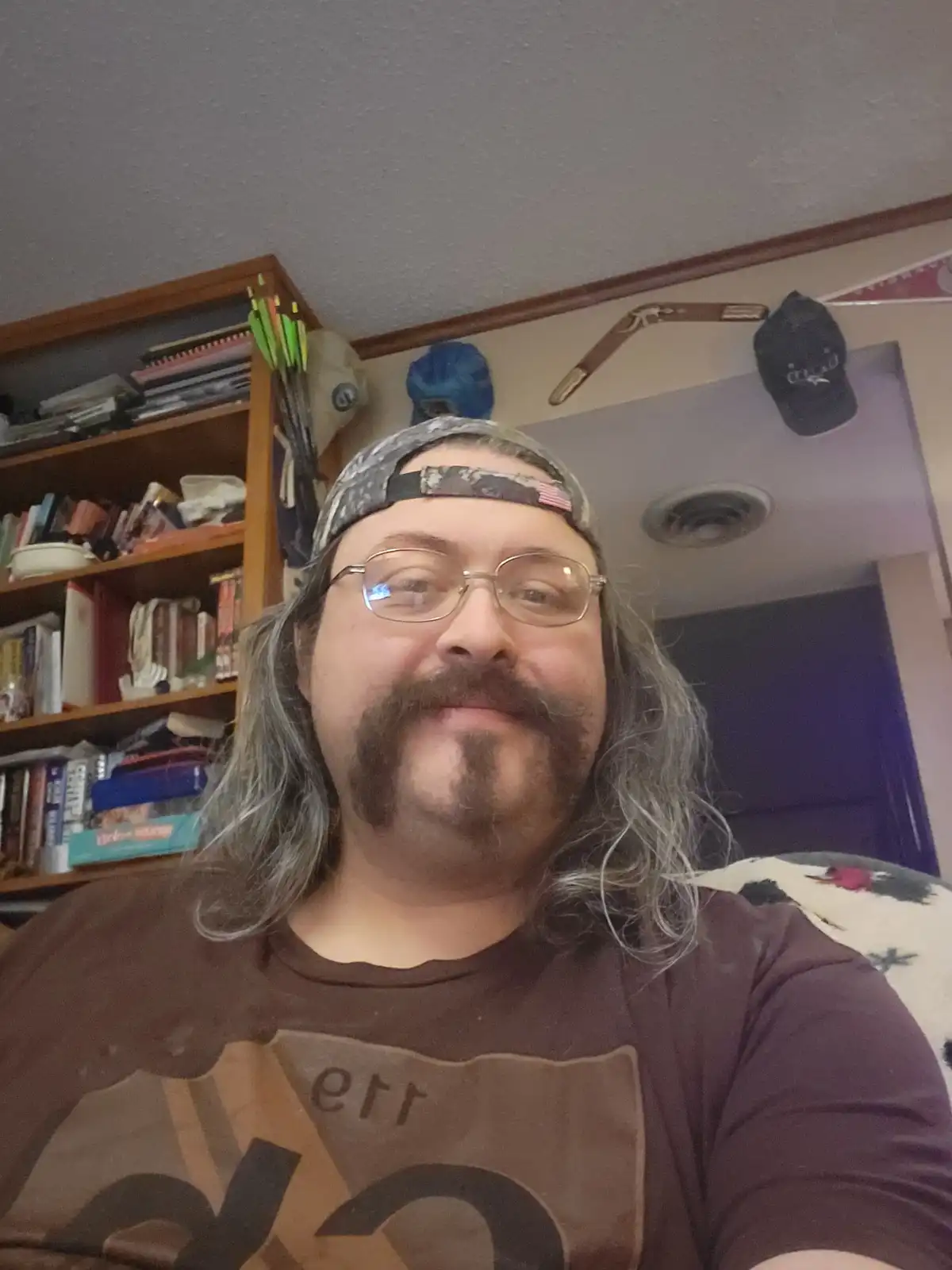 A man with long gray hair, glasses, and a goatee wearing a backward cap and a brown t-shirt takes a selfie. Behind him, a…