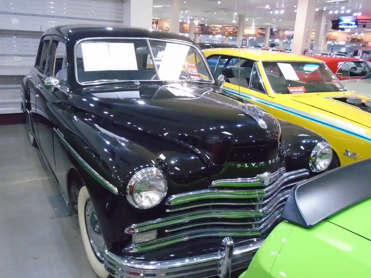 A black 1950s Plymouth sedan is displayed in a car showroom, with a red 'please do not touch' sign visible on its…