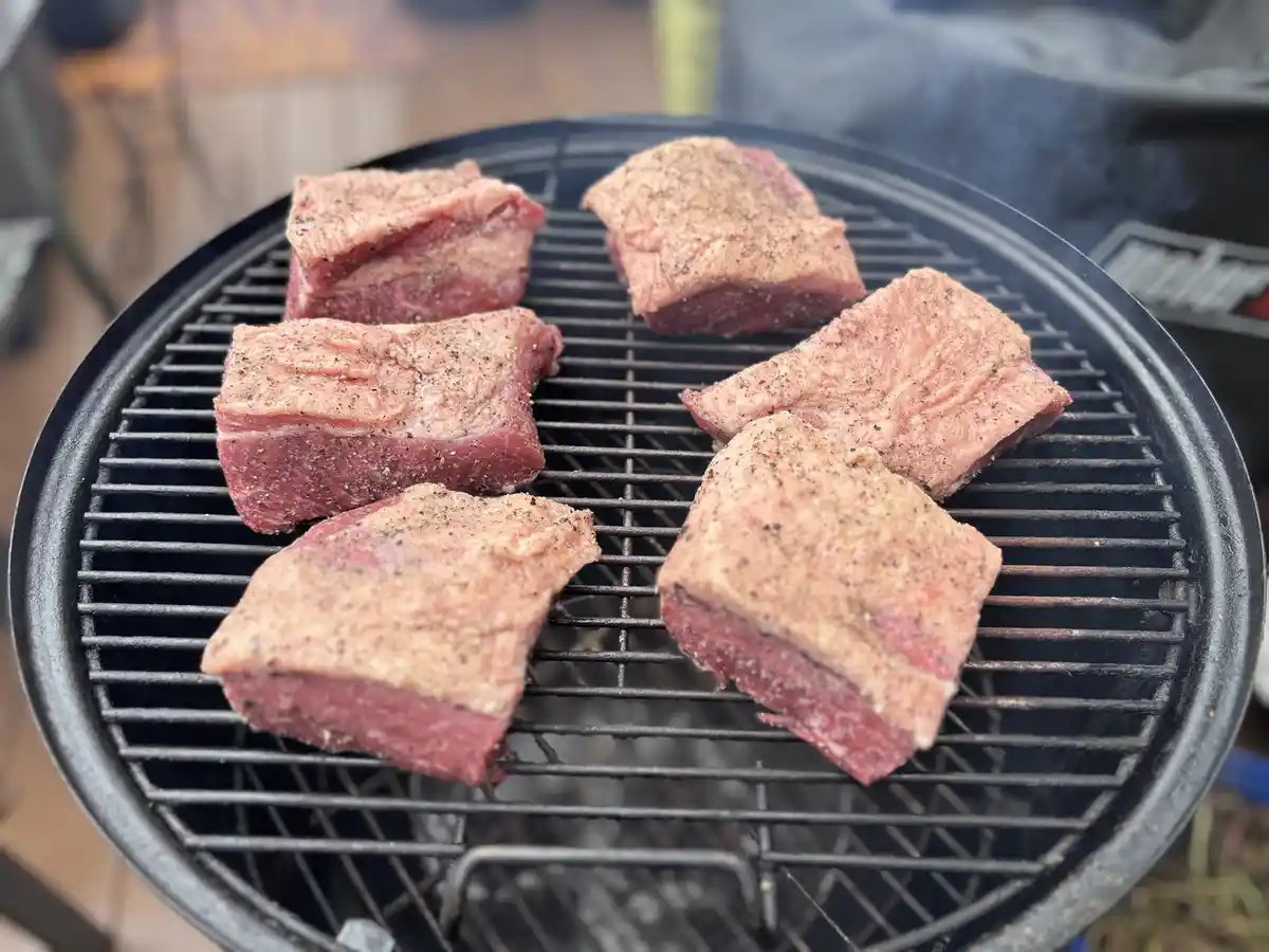 Six raw, seasoned beef steaks are arranged on a black charcoal grill grate, with smoke rising from below. The overlaid text…