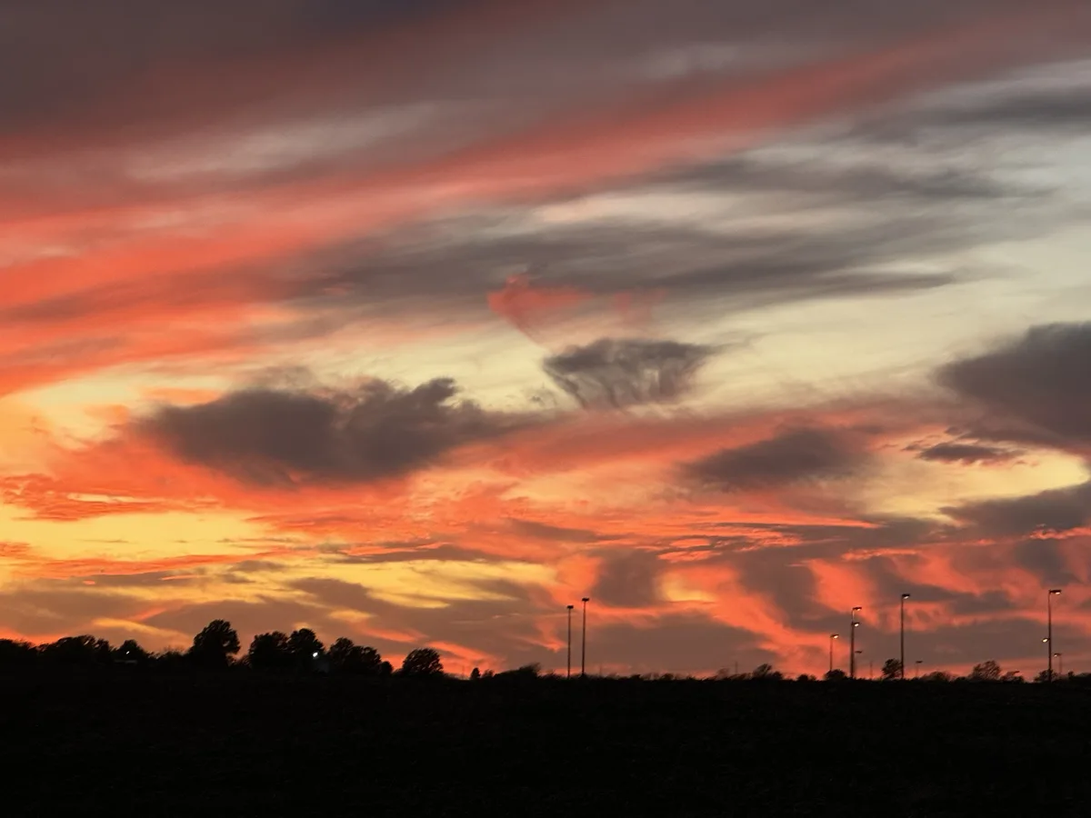 A dramatic sunset with fiery orange, red, and yellow clouds against a dark sky, silhouetting trees and tall light poles on…